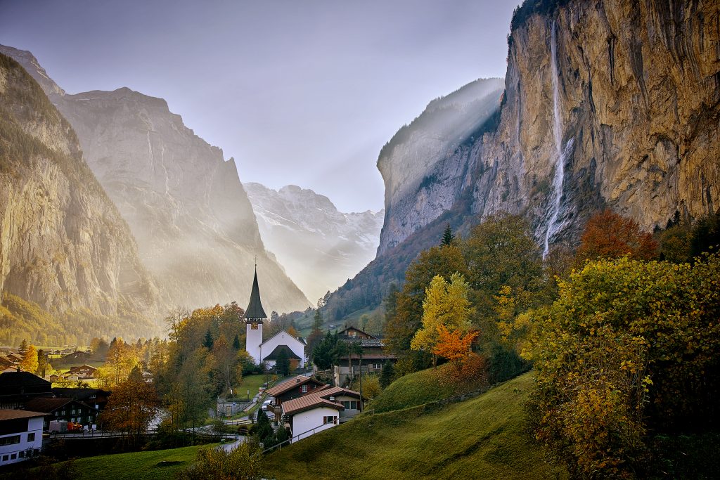 Sun shines across the scenic mountain village of Lauterbrunnen, Switzerland in autumn with the Staubbach waterfall, the Swiss alps, vibrant trees, a small church and a stunning landscape.