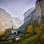 Sun shines across the scenic mountain village of Lauterbrunnen, Switzerland in autumn with the Staubbach waterfall, the Swiss alps, vibrant trees, a small church and a stunning landscape.
